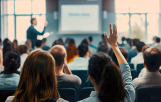 Backs of people in auditorium with presenter at front, woman with hand raised