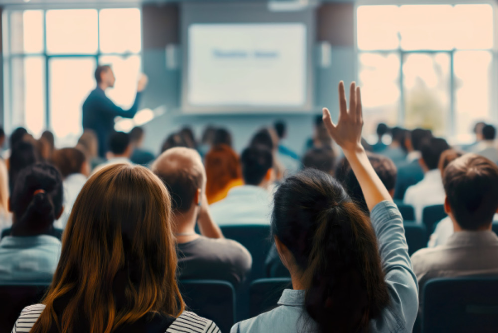 Backs of people in auditorium with presenter at front, woman with hand raised